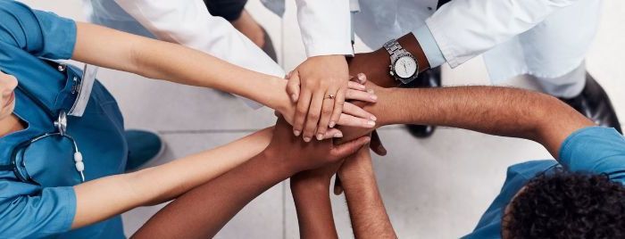 5 Personen stehen im Kreis, sich zugewandt, die Arme sind sternförmig zueinander ausgestreckt und die Hände übereinandergelegt.